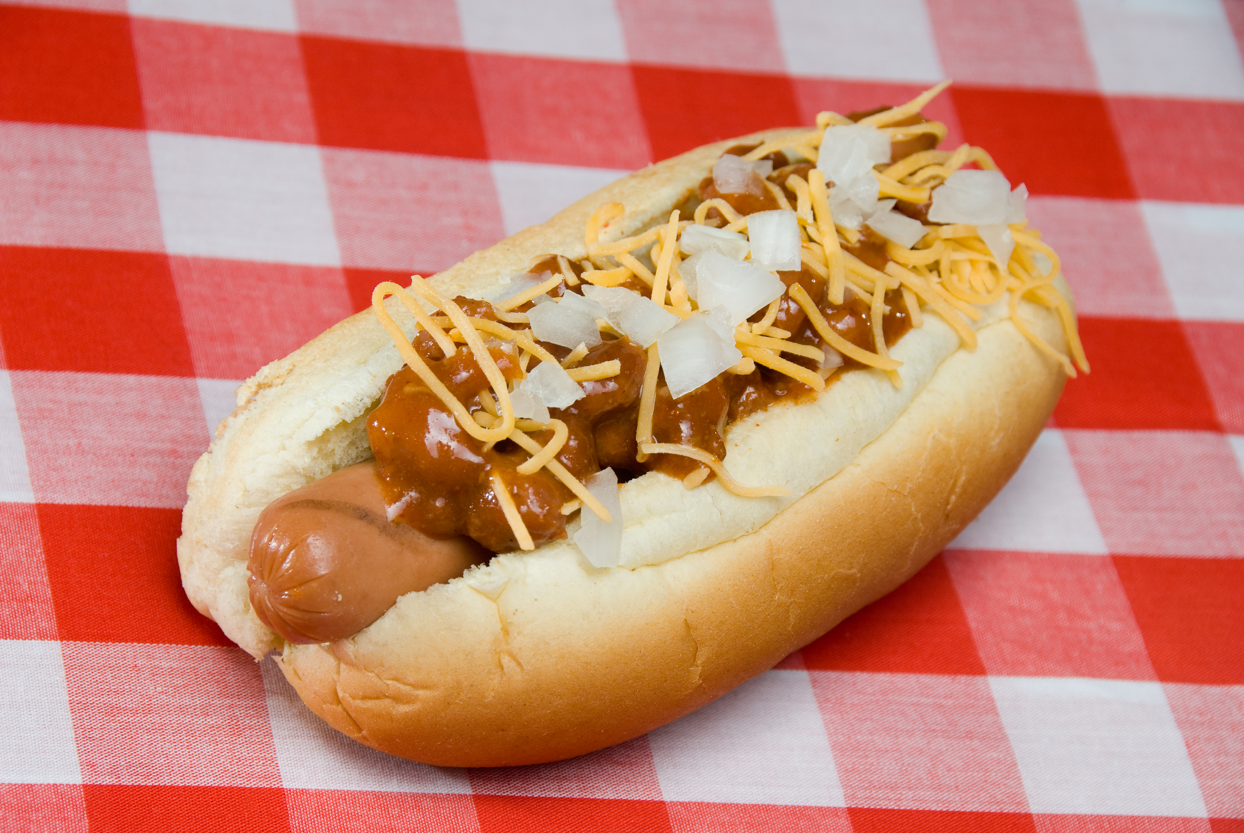 A scrumptious barbecued chili dog with onions, mustard and cheese rests on a picnic table waiting to be consumed.